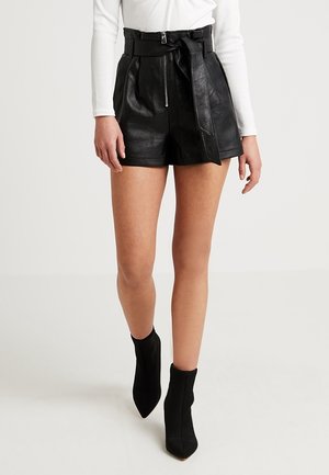Vrouw die zwarte leren shorts met hoge taille en riem draagt, een wit top met lange mouwen en zwarte enkellaarzen met puntige neus, lopend naar voren.