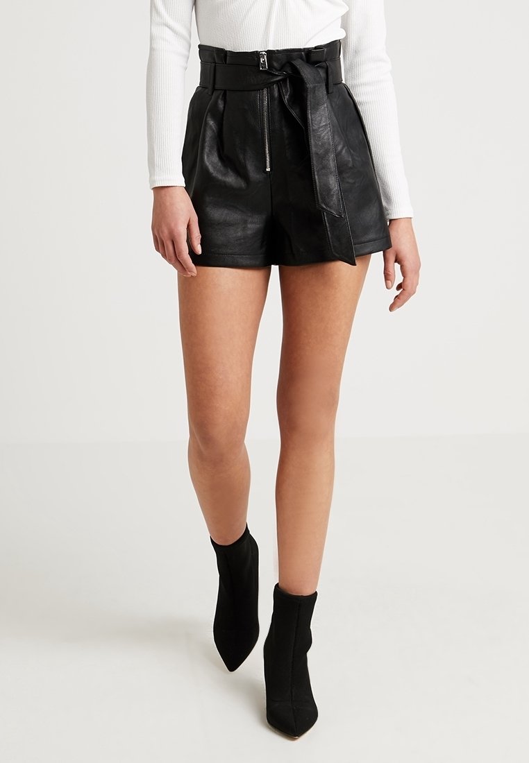 Femme portant un short en cuir noir taille haute avec ceinture, un haut blanc à manches longues et des bottines noires à bout pointu, marchant vers l'avant.