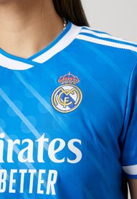 Maglia sportiva blu con accenti bianchi, strisce diagonali e il logo del Real Madrid con una corona, realizzata in tessuto traspirante.