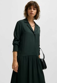 Dunkelgrünes, langärmeliges Kleid mit kragenförmigem Halsauschnitt und einem Knopf. Ausgestellter, geraffter Saum aus glattem Stoff; kombiniert mit einer schwarzen Tasche.
