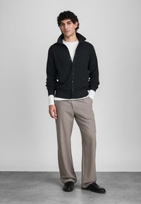 Jonge man die met handen in zijn zakken staat, gekleed in een zwarte knopen cardigan over een witte blouse, beige broek en zwarte schoenen, tegen een grijze achtergrond.