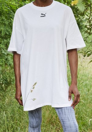 Osoba nosząca oversize białą koszulkę Puma i wzorzyste legginsy, stojąca na zewnątrz z zielenią w tle.