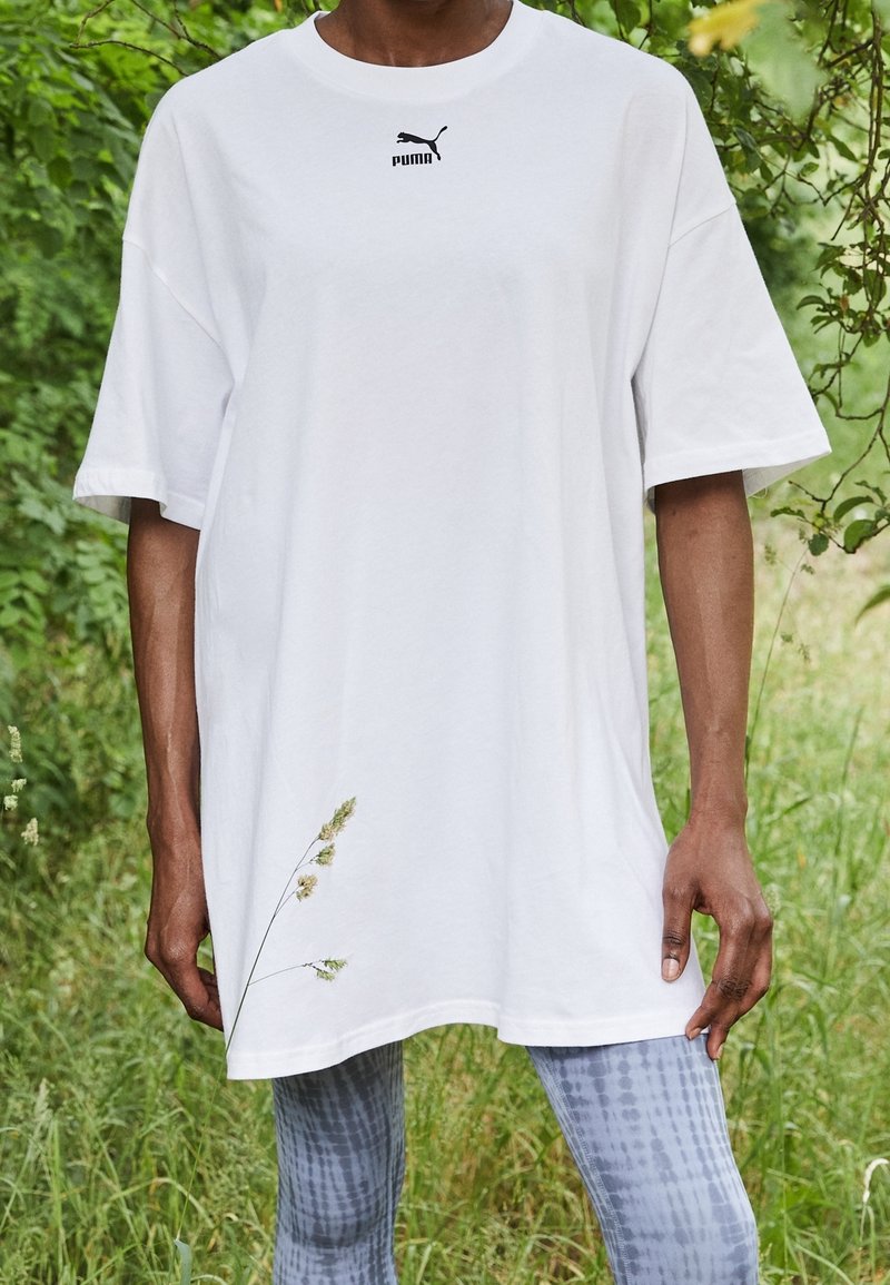 Personne portant un T-shirt blanc Puma surdimensionné et des leggings à motifs, debout en plein air avec de la verdure en arrière-plan.