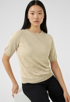 Beige kortærmet striktrøje med rund halsudskæring og ribbet kant; glat tekstur, figursyet silhouet, båret med sorte bukser.