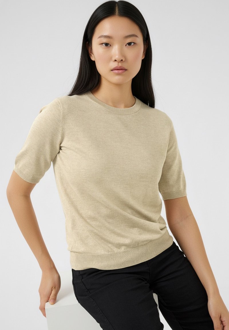 Beige kortærmet striktrøje med rund halsudskæring og ribbet kant; glat tekstur, figursyet silhouet, båret med sorte bukser.