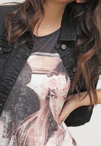 Czarna dżinsowa kurtka noszona na graficznej koszulce z wyblakłym wizerunkiem osoby z kwiatami we włosach, podkreślająca miękką teksturę i stonowane kolory.