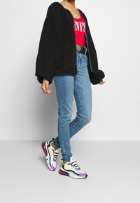 Black faux fur jacket, red tank top with white text, light blue skinny jeans, multicolored sneakers with a wavy design and black accents.