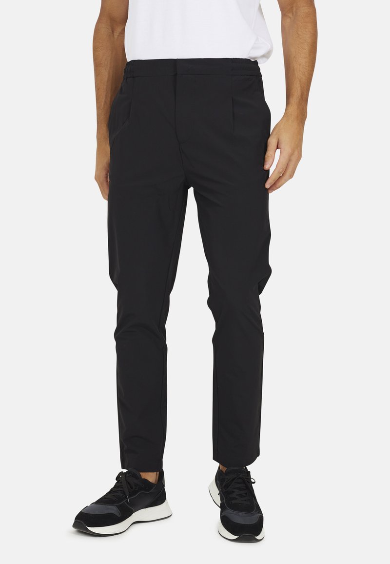 Zwarte, lichte broek met een elastische tailleband, getailleerde pasvorm, zijzakken en gestikte details. Gdragen met zwarte sportieve schoenen.