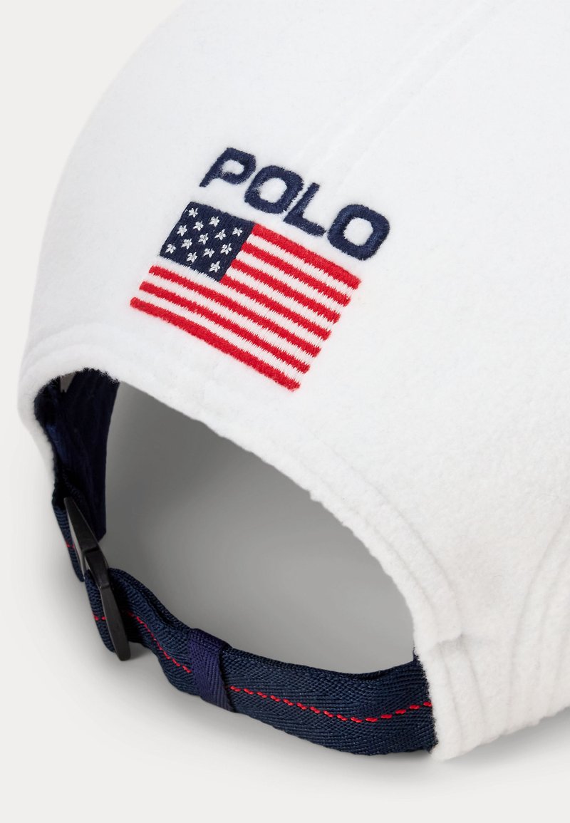 Vit fleecekeps med blå "POLO"-bokstäver och en amerikansk flaggpatch. Den har en marinblå justerbar rem med röda sömmar.