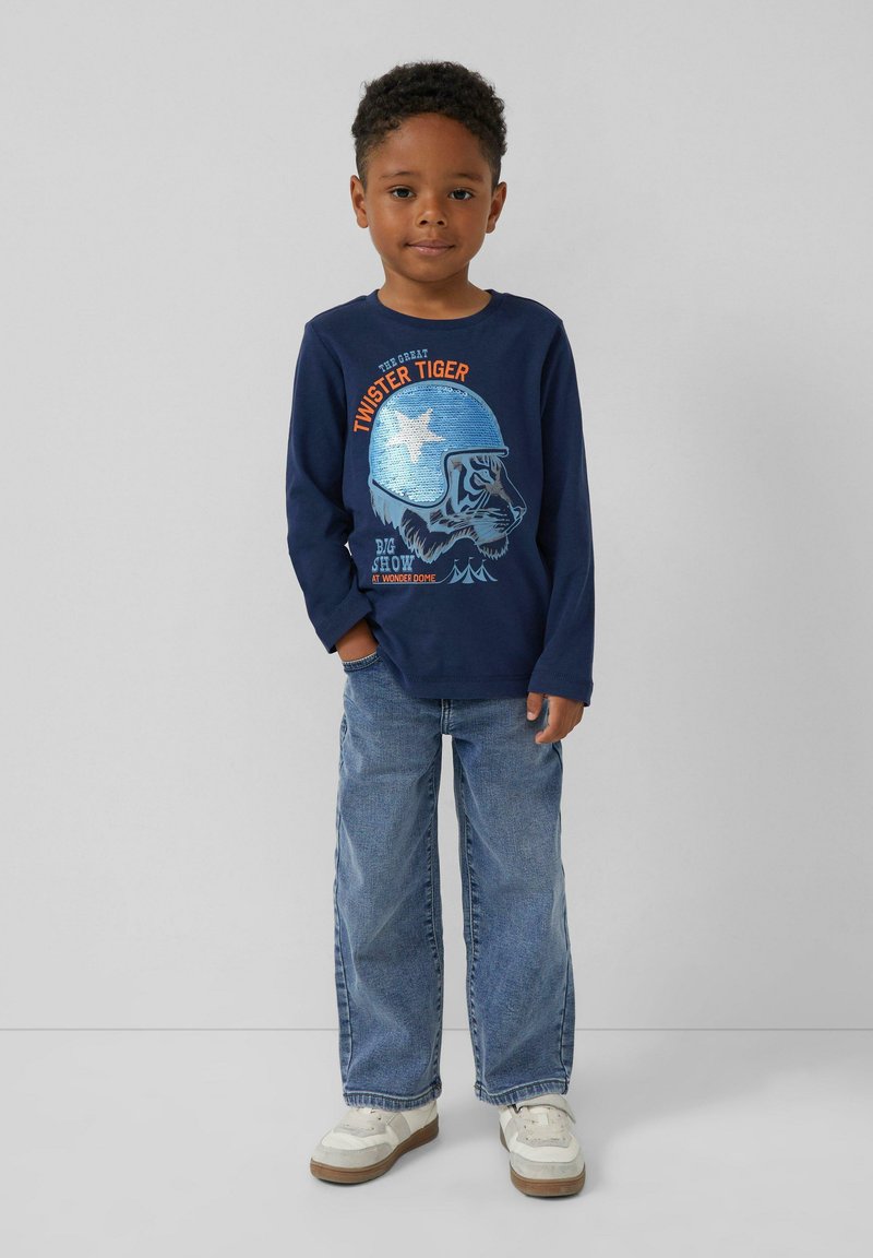Granatowa koszulka z długim rękawem z grafiką tygrysa w niebieskim hełmie, zestawiona z luźnymi dżinsami i białymi trampkami.