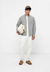 Camicia a righe blu e gialle con bottoni, T-shirt bianca, pantaloni bianchi, tiene in mano una borsa beige, indossa un berretto a quadri marrone e scarpe marroni.