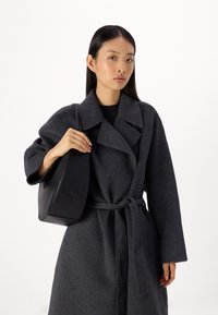 Cappotto grigio di lana con cintura, con ampi rever a incasso. La persona tiene una borsa a mano nera in pelle con una superficie liscia.