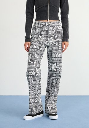 Personne portant un pantalon à motif noir et blanc, une veste courte noire zippée et des chaussures noires à enfiler avec des semelles blanches.