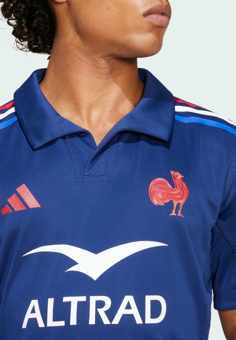Maillot de sport bleu au design à col. Présente un emblème de coq rouge, un logo blanc et des rayures rouges/belges sur les épaules.