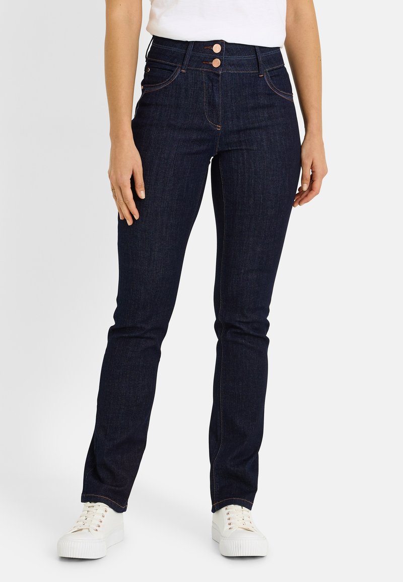 Vaqueros de mezclilla oscura con cintura alta, con dos cierres de botón y costuras de cobre. Combinados con zapatillas blancas.