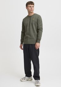 Olive green sweatshirt with a crew neckline, ribbed cuffs, and a small yellow logo on the chest. Paired with black pants and gray sneakers.