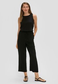 Zwarte mouwloze top gecombineerd met zwarte, loszittende broek met een elastische tailleband en trekkoord. Zwarte platte sandalen maken de outfit compleet.