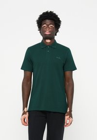 Polo de cor verde escura, feito de tecido texturizado, com uma pala de três botões e um pequeno logótipo bordado no lado esquerdo do peito.