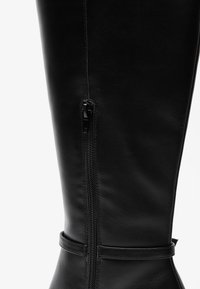 Black leather knee-high boot with a side zipper and strap detail at the ankle. Smooth texture, sleek design, and fitted shape.