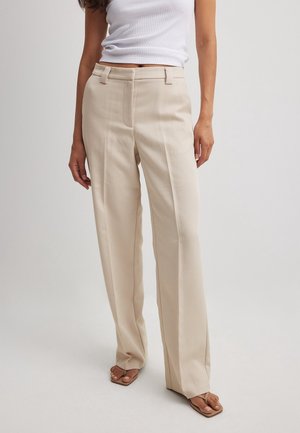 Kobieta w beżowych spodniach z szerokimi nogawkami i wysokim stanem, białym bezrękawnym topie z prążkowanego materiału oraz brązowych sandałach z kwadratowym noskiem na jednolitym tle.