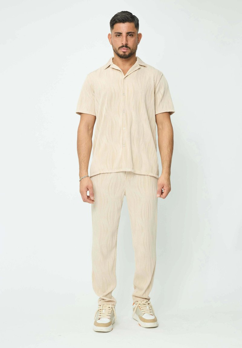 Ensemble beige en deux pièces comprenant une chemise à manches courtes à boutons avec un motif ondulé et un pantalon fuselé assorti. Baskets avec des accents blancs et beiges.