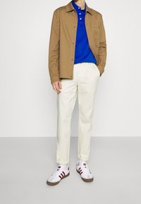 Brun knapjakke over en blå polo skjorte, parret med hvide bukser og hvide sneakers med røde detaljer og brune såler.