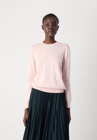 Lys rosa strikkegenser med rund hals og lange ermer, paret med en mørkegrønn plissert skjørt. Har en liten logodetalj på brystet.