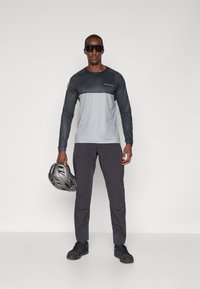 Langärmliges, graues und schwarzes Sportshirt mit kontrastierendem Einsatz, kombiniert mit dunkelgrauen Hosen. Das Model hält einen grauen Fahrradhelm.