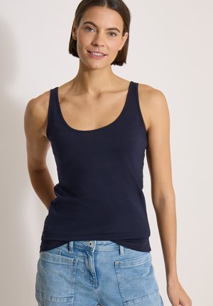 Femme aux cheveux bruns courts portant un débardeur noir et un jean bleu clair, debout devant un fond clair uni.