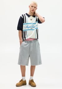 T-Shirt mit kurzen Ärmeln in Blau-weißen horizontalen Streifen, kombiniert mit grauen Shorts und braunen Schnürschuhen. Der Look verkörpert einen lässigen Streetwear-Stil.