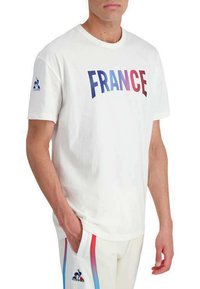 T-shirt en coton blanc avec "FRANCE" écrit en lettres dégradées rouge et bleu. Manches courtes avec un logo sur la manche gauche. Design simple et décontracté.