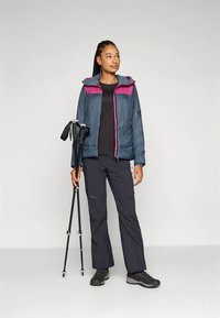 Pikowana kurtka w ciemnym granacie z różowym kapturem, zestawiona z czarnymi spodniami i butami. Użytkownik trzyma kije trekkingowe, stoi w neutralnej pozie.