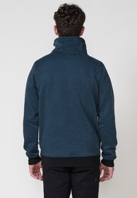 Jersey azul con un patrón, cuello alto, tela texturizada y puños y dobladillo de canalé negros. Costuras detalladas visibles en la parte posterior.