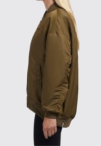 Vue latérale d'une personne portant une veste bomber oversize vert olive avec des poignets et un ourlet côtelés, associée à un pantalon noir.