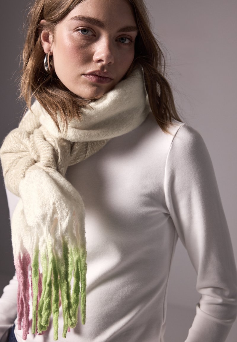 Sciarpa in maglia crema con frange sfumate in rosa e verde, morbida al tatto, indossata in modo sciolto attorno al collo, abbinata a una camicia bianca a maniche lunghe.