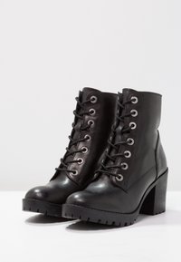 Botas de tobillo de cuero negro con punta redondeada, cordones en la parte delantera, ojales de metal y un tacón grueso. Suela de goma texturizada y con dibujo.