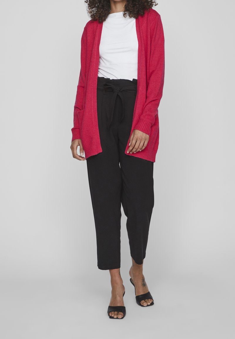 Femme portant un cardigan rouge, un haut blanc, un pantalon noir taille haute avec ceinture et des talons ouverts noirs, debout sur un fond gris.