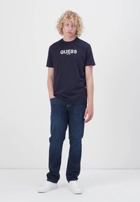 Námornícky modré bavlnené tričko s logom "GUESS" v bielej a šedej farbe, v kombinácii s tmavými džínsami a bielymi teniskami. Voľný strih.