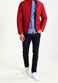 Czerwona pikowana kurtka z zamkiem błyskawicznym z przodu, niebieska koszula z guzikami pod spodem, ciemne dżinsy i białe sneakersy, na tle neutralnym.
