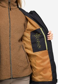 Chaqueta de pelaje sintético marrón con cremallera en el frente, que cuenta con un interior azul marino y iconos amarillos que indican propiedades impermeables, transpirables y a prueba de viento.