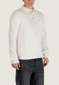 Biały dzianinowy sweter z kołnierzykiem, z trzema guzikami przy dekolcie. Żebrowane mankiety i dół, noszony na ciemnych dżinsach. Logo na piersi.