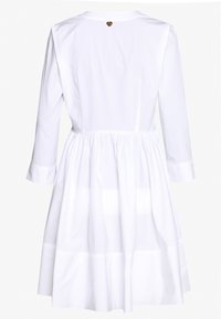 Vestido de algodón blanco con mangas largas, cintura fruncida y falda acampanada. Presenta un pequeño logo de corazón en la parte posterior del escote.