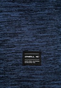 Tissu texturé bleu marine avec un motif subtil. Présente une étiquette O'Neill noire avec un texte blanc, indiquant "Santa Cruz, Californie, Établi en 1952."