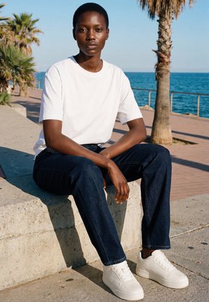 Junge Person mit kurzen Haaren, die ein weißes Hemd, dunkle Jeans und weiße Turnschuhe trägt und auf einer Betonmauer neben Palmen und dem Ozean sitzt.