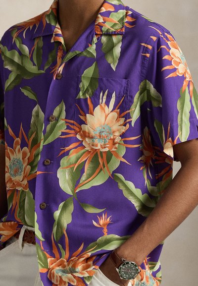 Mężczyzna ubrany w purpurową, krótkorękawową koszulę na guziki z dużym pomarańczowo-zielonym kwiatowym wzorem, białe spodnie i zegarek na nadgarstku, ręka w kieszeni.