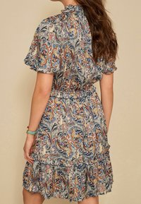 Femme aux longs cheveux bruns portant une robe à manches courtes avec un imprimé paisley multicolore et un ourlet festonné, de dos contre un fond beige.
