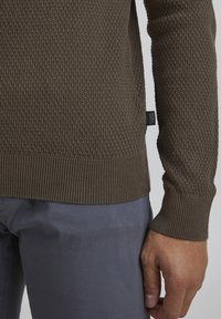 Brown textured knit sweater with ribbed cuffs and hem, featuring a woven pattern. Tag visible on the side near the bottom hem.