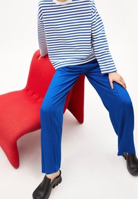 Modré široké kalhoty z elastického materiálu, v kombinaci s bílou a modrou pruhovanou košili s dlouhým rukávem, nošeny s černými dřeváky. Červená sochařská židle.