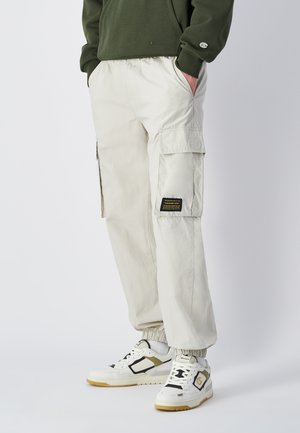 Lichtbeige cargobroek met elastische boorden, meerdere zakken en een zwart patchdetail. G gedragen met wit-zwart sportieve schoenen.