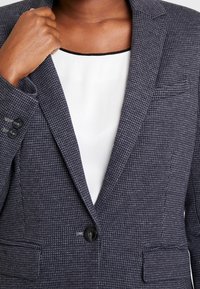 Persona che regola il colletto di un blazer grigio scuro a trama sopra a una maglietta bianca con il bordo del collo nero.